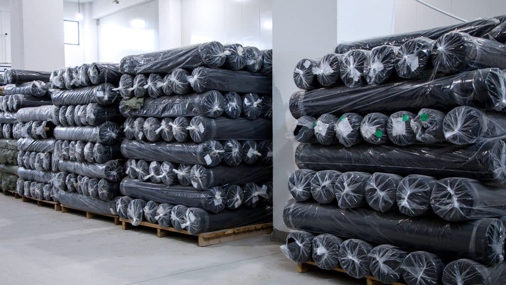 Neatly stacked Black and D. Grey performance fabric rolls in a bright activewear manufacturing warehouse, reserved for hybrid seamless apparel production.